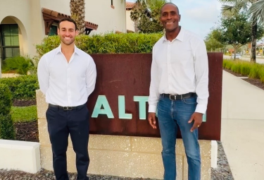 Two men in white shirts and dark pants posing beside a brown sign with the letters 'ALT' visible, outdoors on a sunny day.