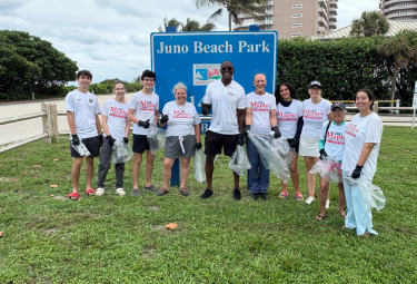 Group of volunteers in white shirts pose in front of the Juno Beach Park sign, each holding a trash bag after a beach cleanup, with palm trees and buildings in the background.