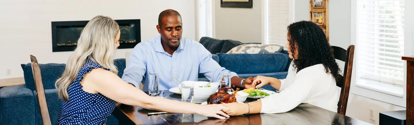 Three adults share a meal around a dining table in a bright living room, with a roasted chicken centerpiece and bowls of food nearby.