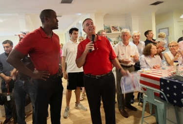 Man in a red polo speaks into a microphone at a crowded indoor gathering with patriotic decor in the background.