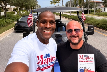 Two men smile for a selfie in a golf cart on a sunny street, wearing campaign shirts (one white Jon Maples shirt, one black shirt) with small American flags nearby.