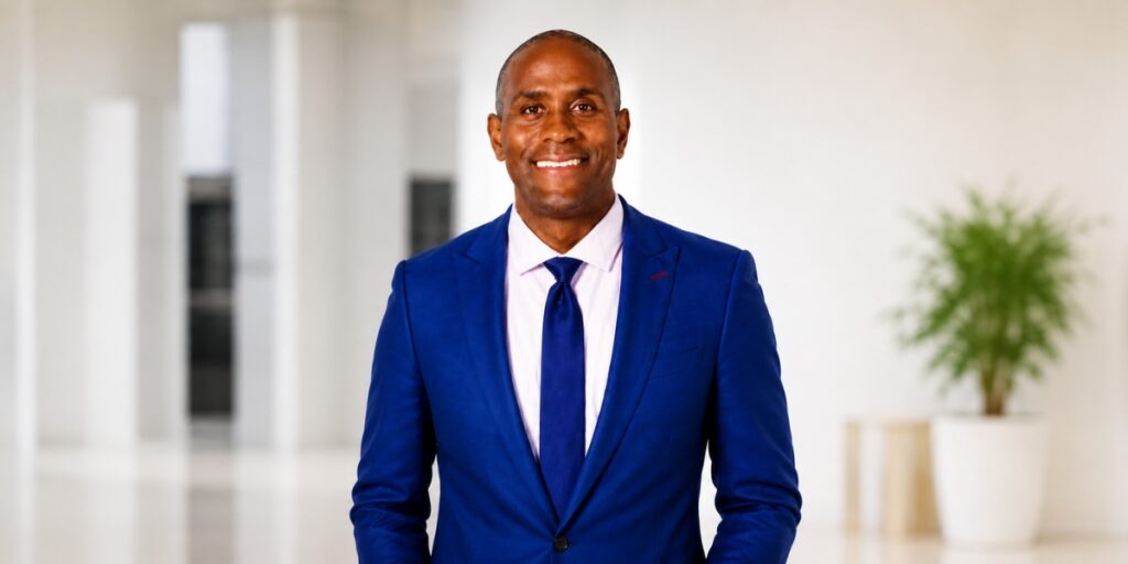Smiling man in a navy blue suit and tie standing in a bright modern office lobby.