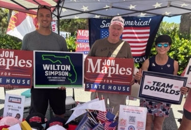 Three volunteers at an outdoor campaign booth hold signs reading Jon Maples for State House, Wilton Simpson, and Team Donalds, with American flags and banners in the background.