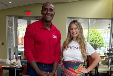Two adults pose for a photo indoors; man in a red polo with a logo stands beside a smiling woman in a white T‑shirt.