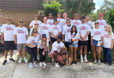Group of about 20 people wearing white t-shirts standing and kneeling outdoors for a charity event, with a brown dog at the front center.