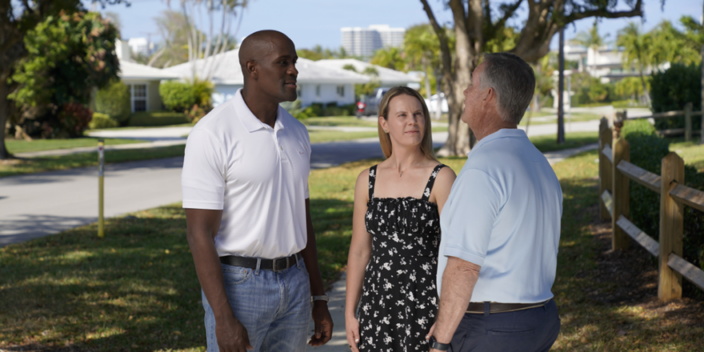 Three adults stand on a sidewalk in a suburban neighborhood, conversing outdoors near a wooden fence and trees on a sunny day.