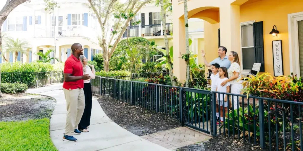 A welcoming scene outside a sunny apartment complex: a couple on a curved walkway greet a family on a nearby porch waving hello from a house with a Welcome sign.