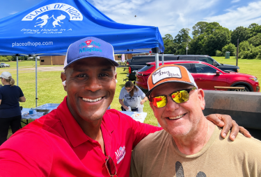 Two men posing and smiling at an outdoor event under a blue Place of Hope tent, one in a red polo and the other in a tan shirt with sunglasses at his eyes.