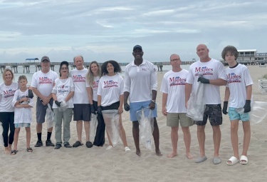 Group of volunteers on a sandy beach in white shirts, holding trash bags after a beach cleanup.