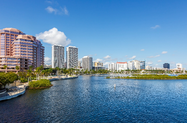 Urban waterfront with high-rise buildings along a calm blue body of water and palm trees by the shore.