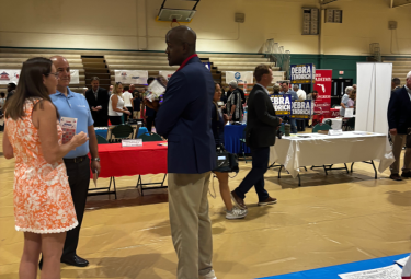Event 51 - votemaples.com People conversing at an indoor career fair with exhibitor booths set up along the gymnasium floor.