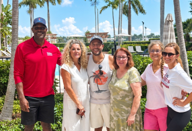 Event 52 - votemaples.com Six people posing together outdoors at a sunny tropical resort with palm trees in the background, smiling for the photo.