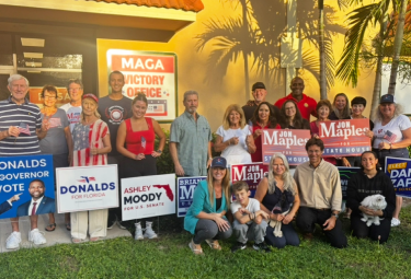 Event 53 - votemaples.com Group of diverse volunteers posing outdoors with campaign signs for Donalds and Ashley Moody, among others, with a tropical backdrop and a red campaign storefront in the background for context.