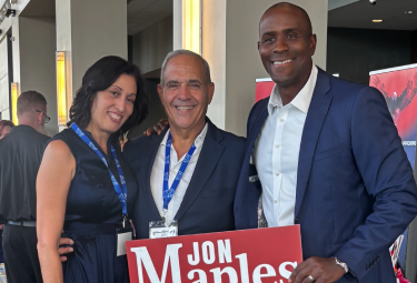 Event 54 - votemaples.com Three adults at a conference posing for a photo, two women and one man in the middle, all wearing event lanyards; the man on the right holds a red-and-white sign with large text (partially visible).