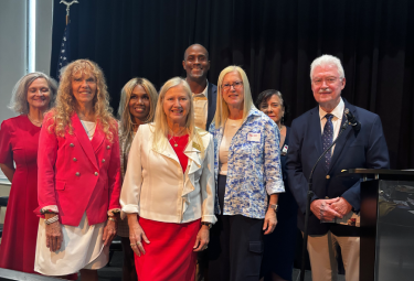 Event 55 - votemaples.com Group of eight adults in business attire posing on stage for a formal event photo.