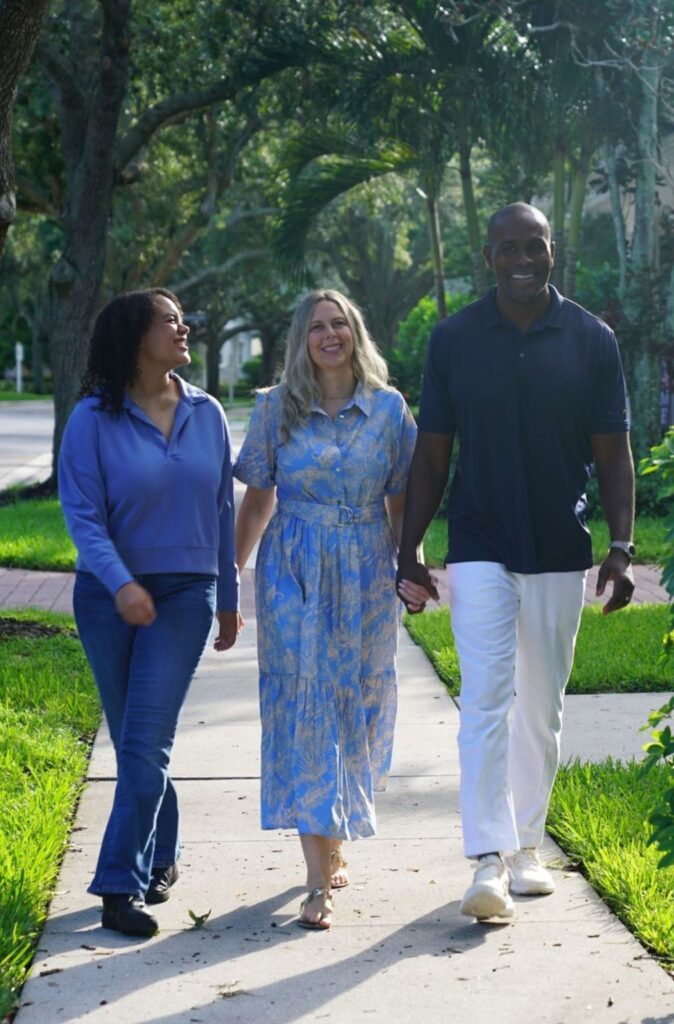 Three adults walk arm-in-arm down a sunny suburban sidewalk, smiling, with green lawns and trees around them.