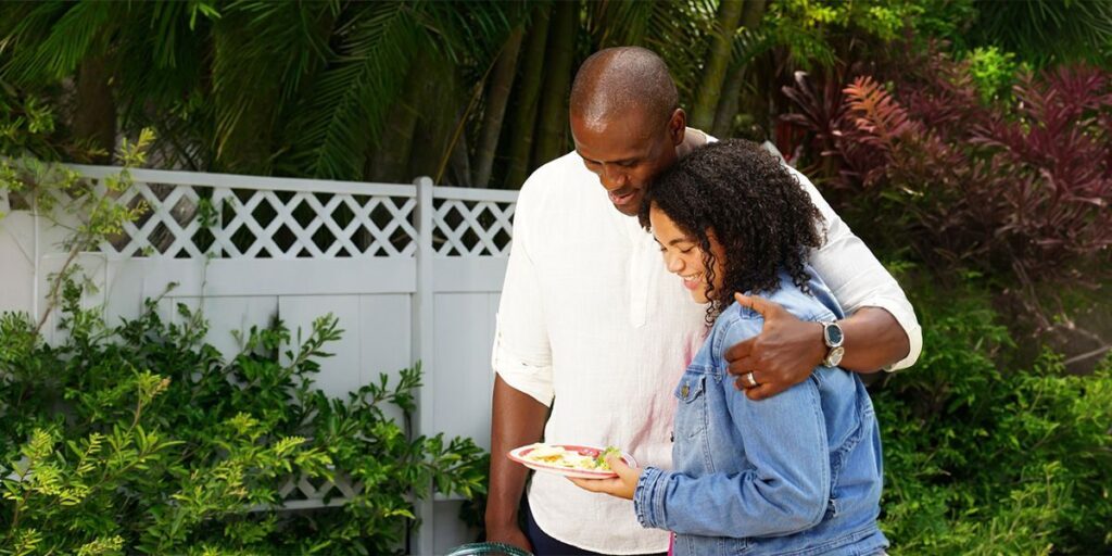 Couple smiling outdoors in a garden, the man embracing the woman while she holds a plate of food.